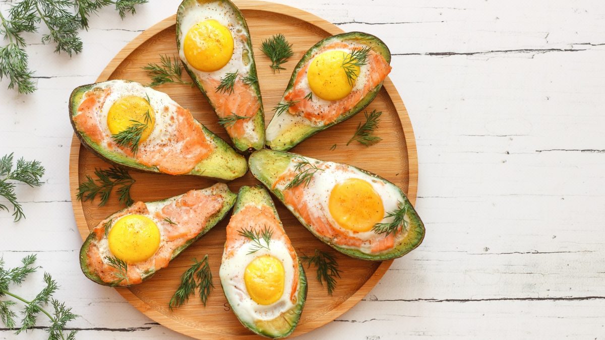 Draufsicht: Sechs gefüllte Avocado mit Lachs und Ei auf einem hellbraunen Teller. Der liegt auf einem weißen Untergrund aus Holz. Drum herum liegen frische Dillspitzen.