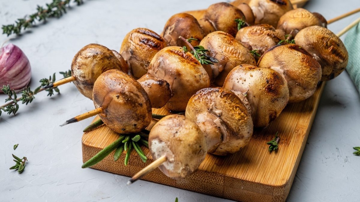 Frontal: Ein Brettchen aus Holz liegt auf einem hellem Untergrund. Auf ihm sind drei marinierte Champignon-Spieße.