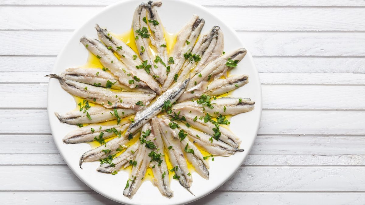 Draufblick: Marinierte Sardellen auf einem weißen, runden Teller. Die Tapas-Platte liegt auf einem weißen Untergrund aus Holz.