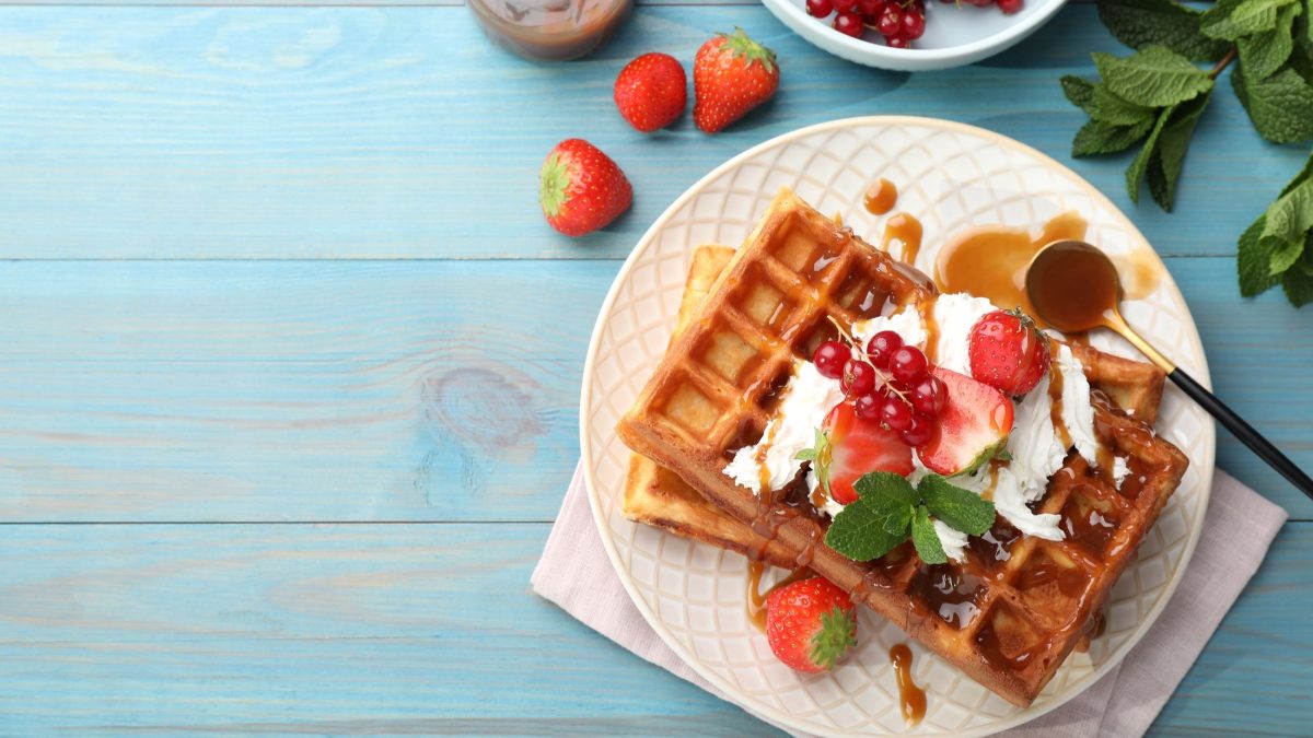 Draufsicht: Ein Teller mit süßen Mutti-Waffeln auf einem weißen Teller. Die Waffeln sind mit Eiscreme und Beeren garniert. Der Teller steht auf einem blauen Untergrund aus Holz. Drum herum liegen frische Beeren.
