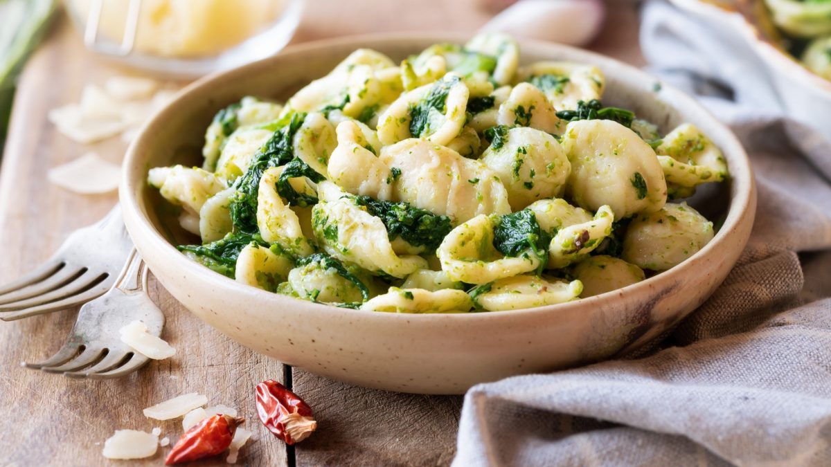 Draufsicht, schräg: Eine hellbraune Schüssel Orecchiette mit Cima di Rapa steht auf einem Holztisch. Drum herum liegen ein Küchenhandtuch, zwei Gabeln sowie eine getrocknete Chili.