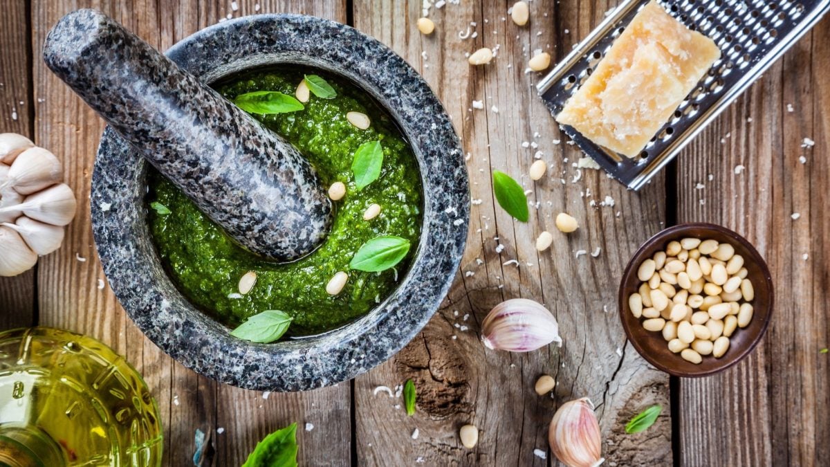 Draufsicht. Auf einem Holztisch liegen alle Geräte und Zutaten um Pesto-Rezepte zuzubereiten. Unter anderem sind dort ein Mörser, Käse, Pinienkerne, Knoblauch und Öl.
