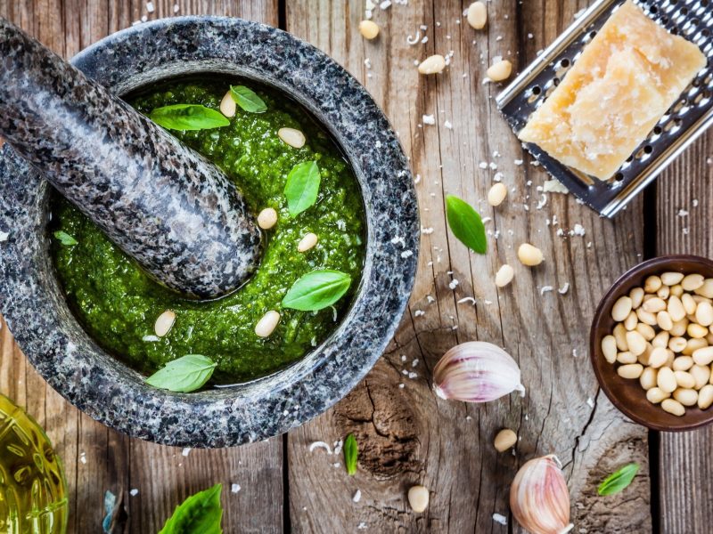Draufsicht. Auf einem Holztisch liegen alle Geräte und Zutaten um Pesto-Rezepte zuzubereiten. Unter anderem sind dort ein Mörser, Käse, Pinienkerne, Knoblauch und Öl.