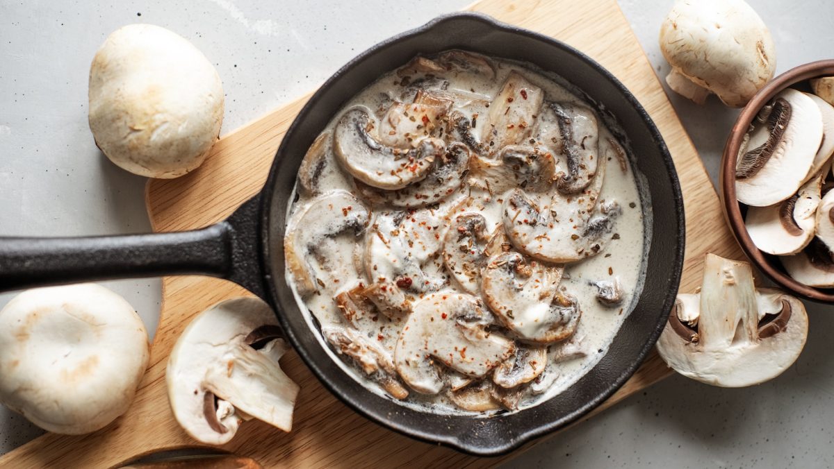 Draufsicht: Auf einem hellem Untergrund liegt eine dunkle Pfanne in der eine Pilz-Sahne-Sauce ist. Die wurde nach dem Grundrezept zubereitet. Drum herum liegen frische Champignons.