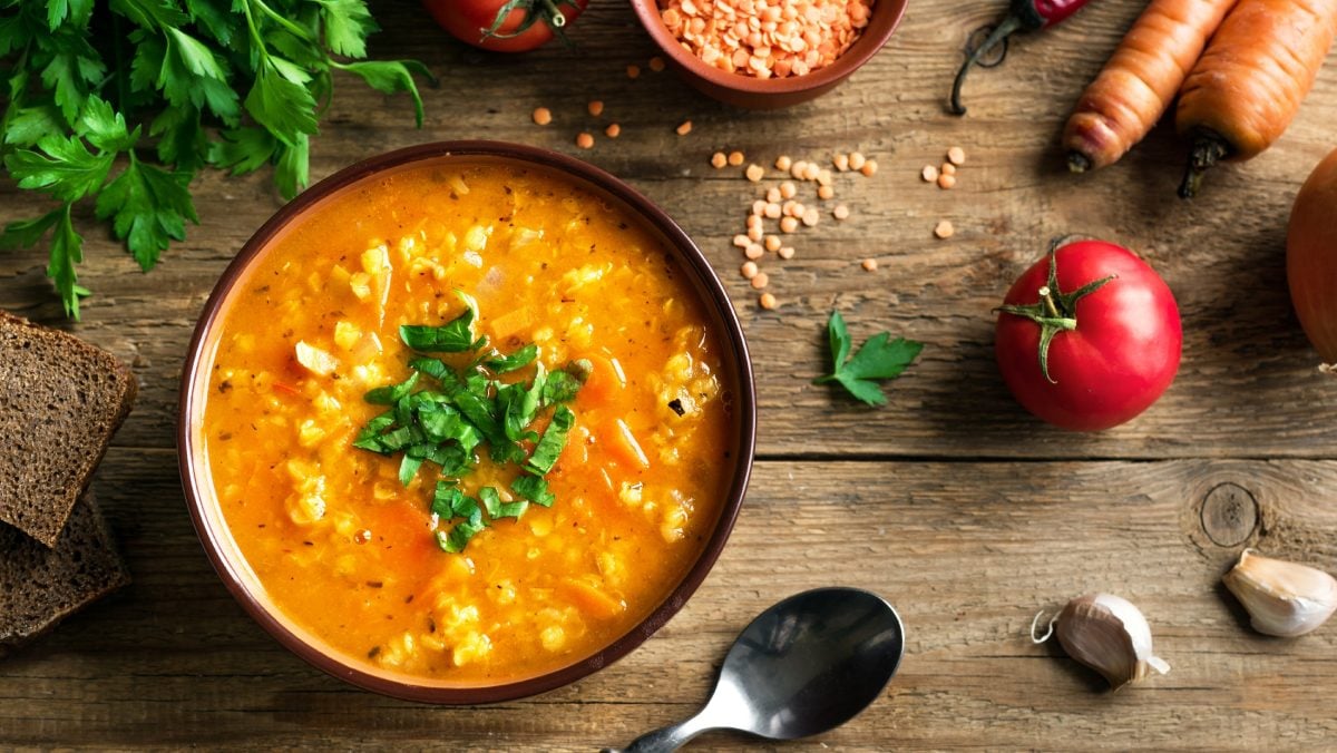 Draufsicht: Eine rustikale Unterlage aus Holz. Auf der stehe ein Teller Rote Linsensuppe mit Bulgur. Drum herum liegen frisches Gemüse und ein Suppenlöffel.