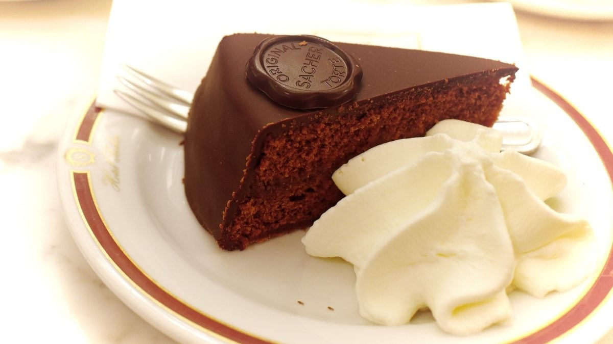 Draufsicht schräg: Ein weißer Teller mit mit einem dünnen roten Rand. Auf dem ist ein Stück Sacher-Torte mit Sahne. Daneben liegt eine Kuchengabel.