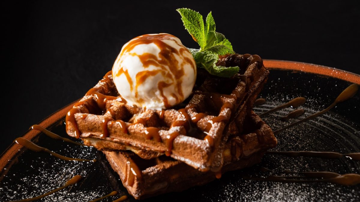 Frontal: Vor dunklem Hintergrund und auf einem dunklem Hintergrund sind zwei Schoko-Eis-Waffeln. Auf denen sind eine Kugel Caramell-EIs und ein Blatt frische Minze.