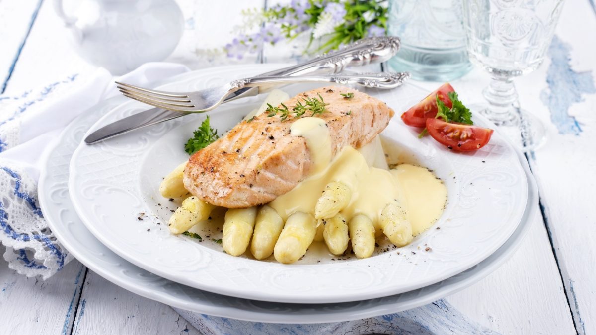 Draufsicht schräg: Ein festlich gedeckter, frühlingshafter Tisch mit einem Teller Spargel mit Lachs und Hollandaise. Am Tellerrand liegen zwei kleine Tomatenstückchen und ein massives, silbernes Besteck.