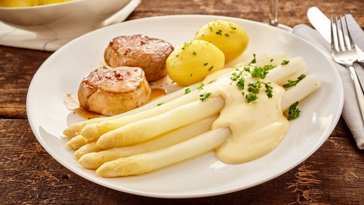 Draufsicht, schräg: Ein weißer Teller mit Spargel mit Medaillons und Sauce hollandaise liegt auf einem Untergrund auf Holz.