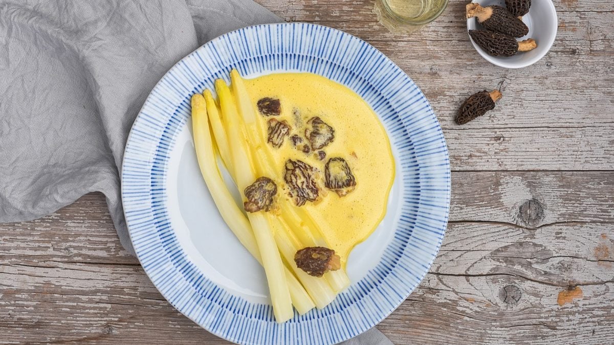 Draufsicht: Ein blau-weißer Teller Spargel mit Morchel-Cremesauce liegt auf einem Holztisch.