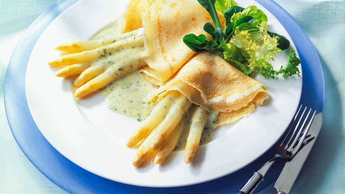 Draufsicht, Ein weißer Teller, der auf einem größeren blauen Teller liegt. Auf dem sind zwei Spargel-Pfannkuchen mit Sauce Hollandaise sowie ein paar Salatblätter. Auf dem blauen Teller liegen ein Messer und eine Gabel.