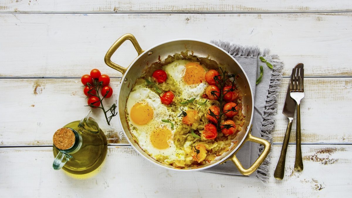 Draufsicht: Eine Pfanne Spiegeleier mit geschmorten Tomaten auf einem weißen Untergrund aus Holz. Daneben liegen Besteck, ein Küchentuch und eine Flasche mit Speiseöl.