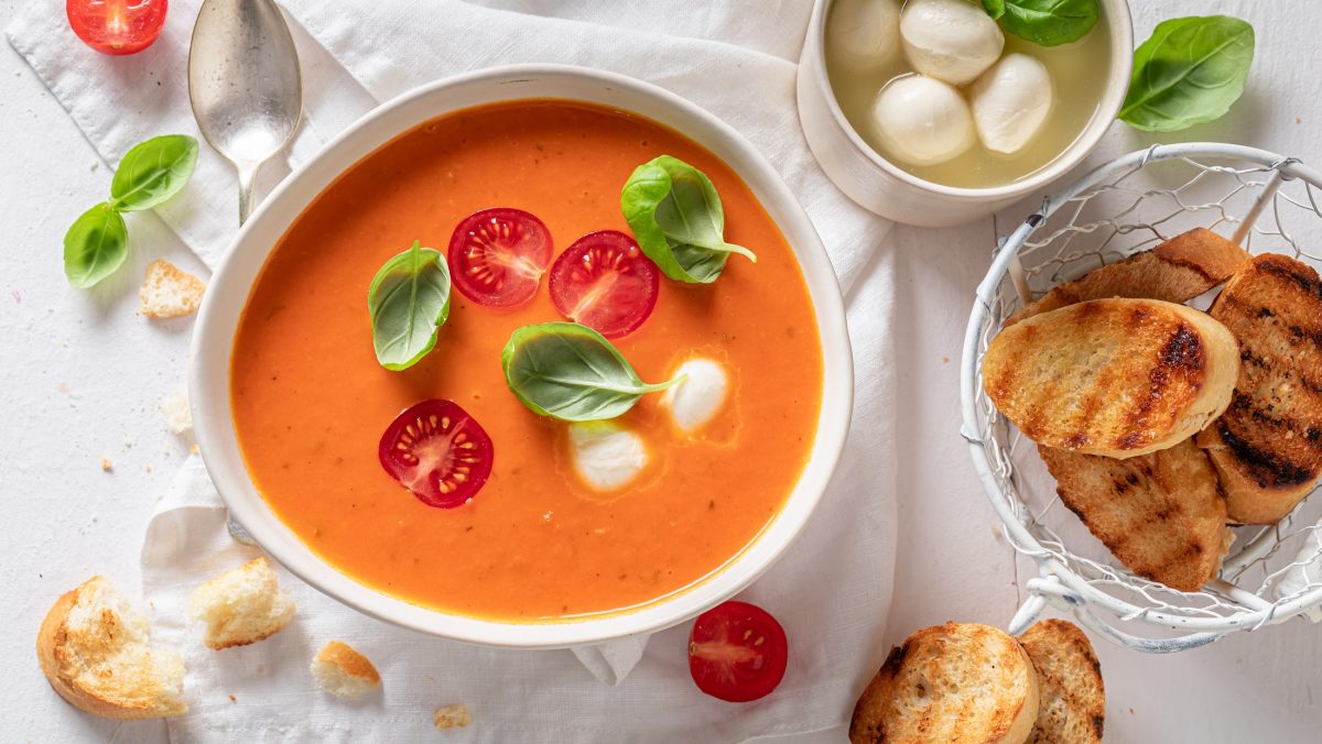 Draufsicht: Ein weißer Untergrund, auf dem eine weiße Schüssel Tomatensuppe alla Caprese steht. Drum herum gibt es eine Schalle mit Mozzarella-Kugeln, frisches Basilikum und geröstete Weissbrotscheiben.