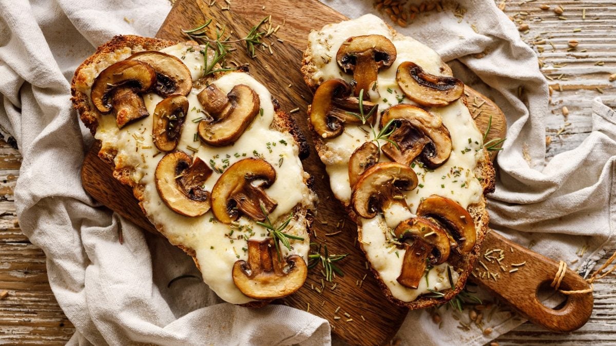 Draufsicht: Ein überbackenes Champignon-Käse-Sandwich liegt auf einem Holztisch. Daneben liegt ein überbackenes Käse-Sandwich mit Pilzen.