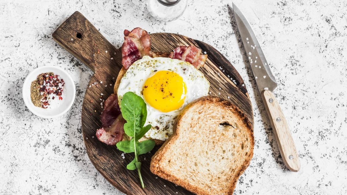 Draufsicht: Ein Uitsmijter met ham liegt auf einem Toast. Es handelt sich um ein gebratenes Spiegelei mit Bacon auf einem Toast, dass auf einem Brettchen auf einem hellen Untergrund liegt.