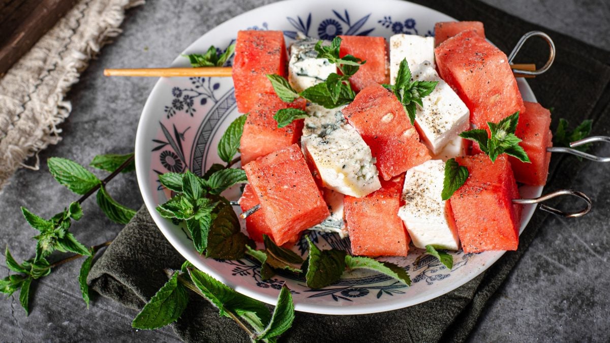 Die Melonen-Feta-Gorgonzola-Spieße auf einem weißen Teller mit blauer Verzierung und frischen Minzzweigen. Alles von oben auf einem grauen Tisch fotografiert.