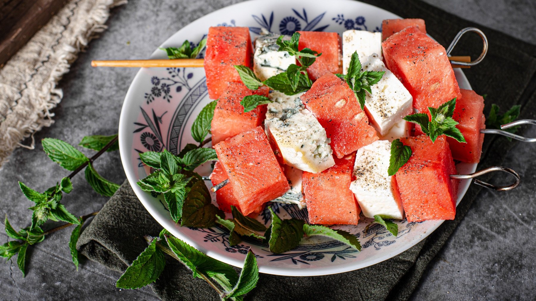 Die Melonen-Feta-Gorgonzola-Spieße auf einem weißen Teller mit blauer Verzierung und frischen Minzzweigen. Alles von oben auf einem grauen Tisch fotografiert.