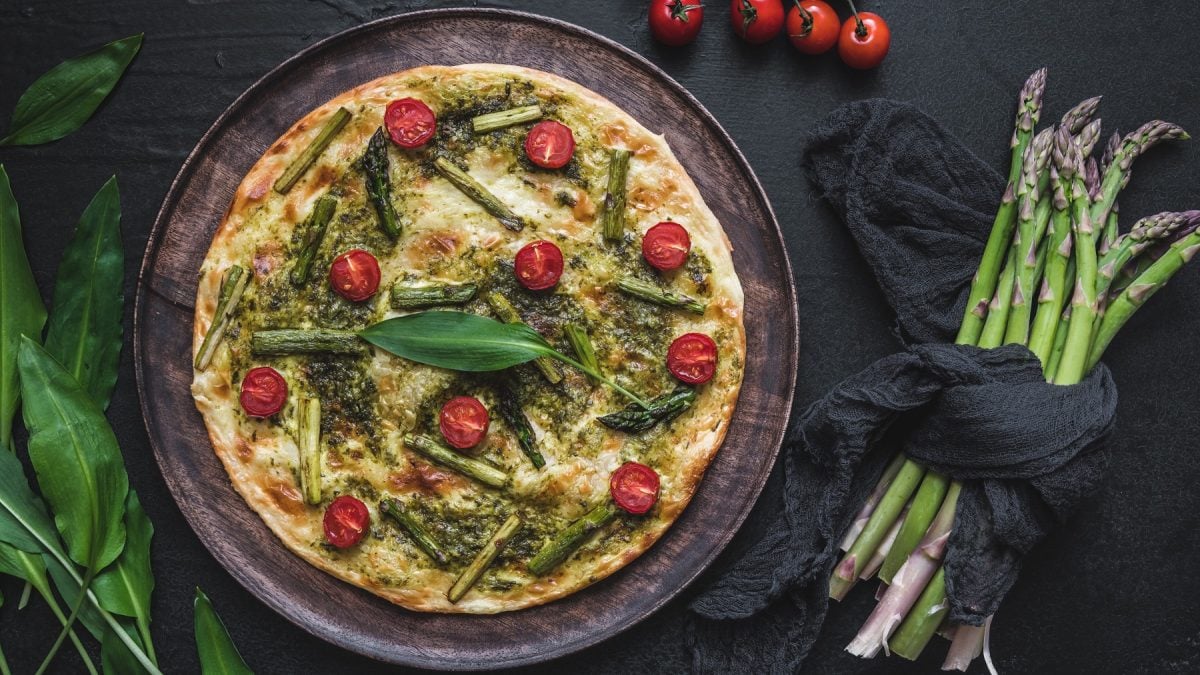 Flammkuchen mit Bärlauch-Pesto, grünem Spargel und Cherry-Tomaten vor dunklem Hintergrund, daneben frischer Spargel und Cherry-Tomaten.