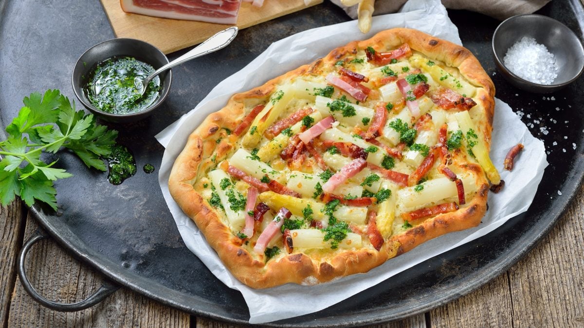 Flammkuchen mit weißem Spargel und Streifen aus Südtiroler Speck. Daneben Schüssel mit Petersilienöl. Alles auf einem schwarzen Tablett.