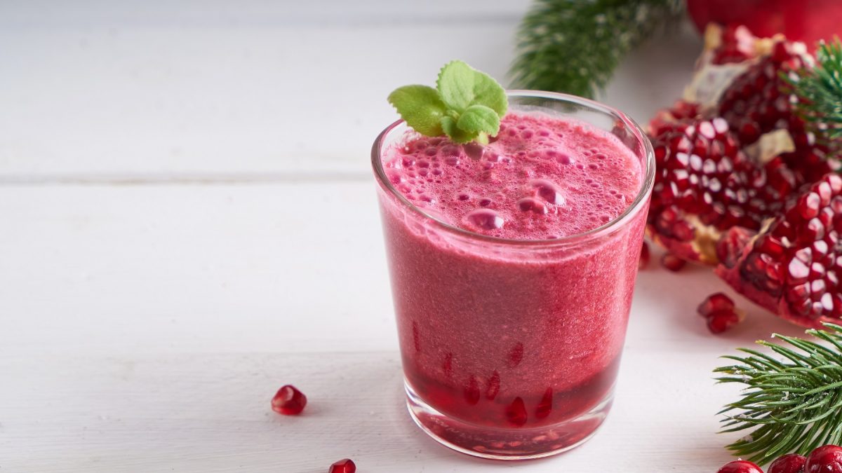 Granatapfel-Smoothie in einem Glas garnier mit frischer Minze, daneben ein angeschnittener Granatapfel.