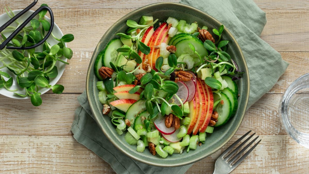 Ein dunkelgrüner Teller mit dem Gurkensalat auf einem Holztisch direkt von oben fotografiert. Darunte reien hellgrüne Serviette und daneben ein paar Sprossen.