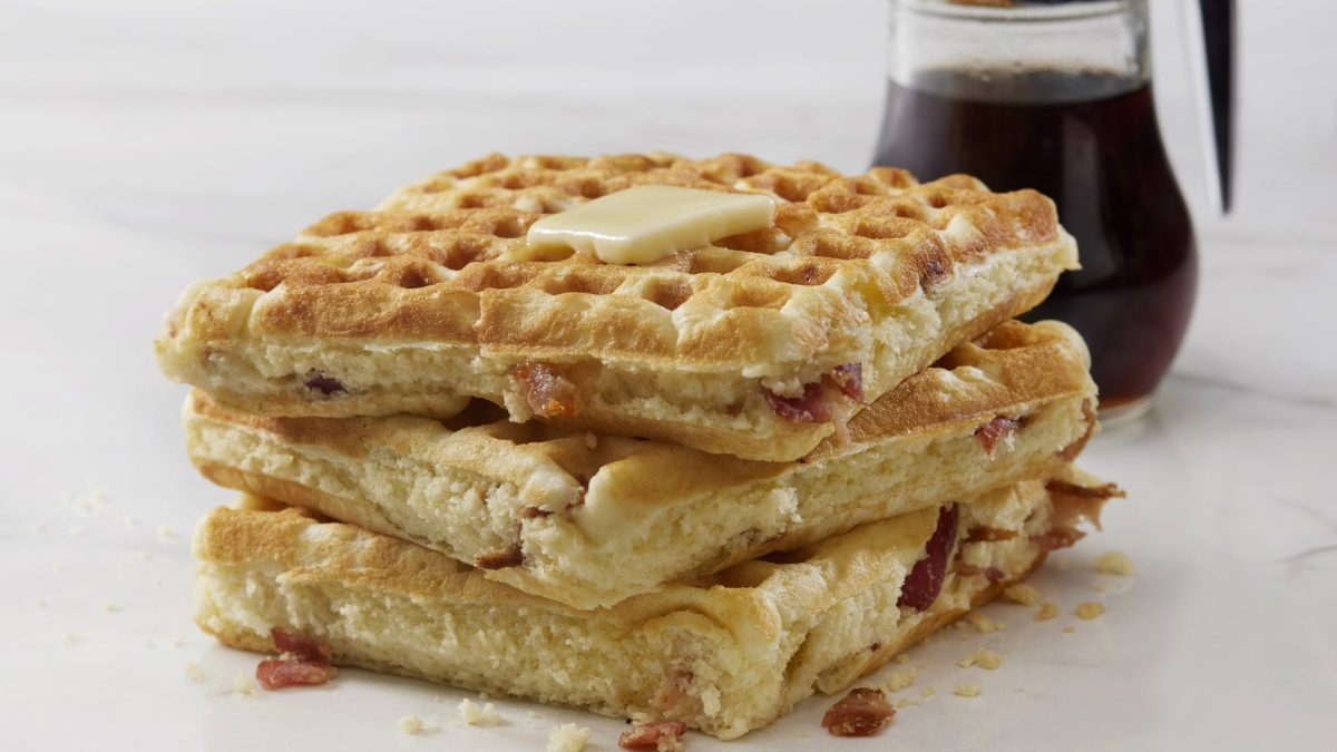 Frisch gebackene herzhafte Waffeln mit schmelzender Butter und Speckstücken, Ahornsirup im Hintergrund.