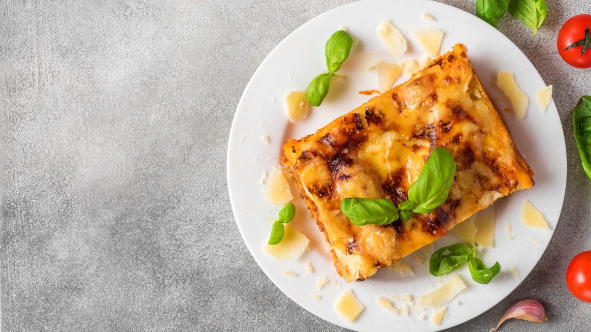 Ein Stück vegane Hummus-Lasagne auf einem weißen Teller. Darauf veganer Parmesan verteilt. Der Hintergrund ist grau.