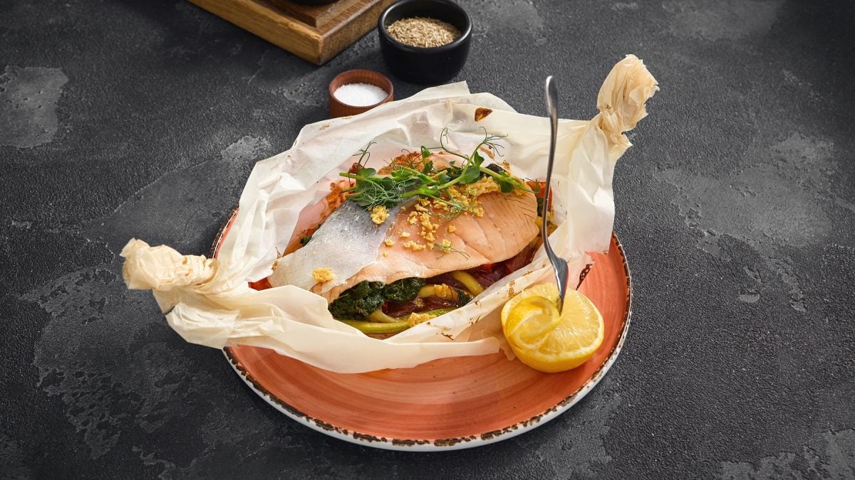Geöffnetes Pergamentpapier auf einem rötlichen Teller mit dem Lachs und Gemüse.