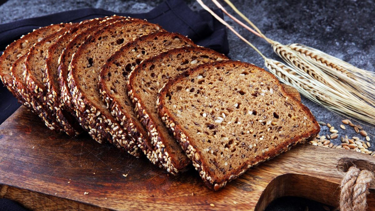 Das angeschnittene Brot auf einem Holzschneidebrett und dunkelblauem Untergrund. Dahinter frisches Getreide. Alles von schräg oben fotografiert.