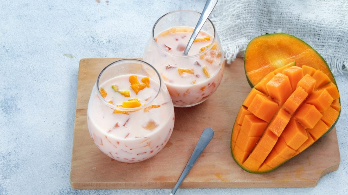 Mango Sago in Gläsern auf einem hölzernen Brett. Rechts auf dem Foto halbe Mangos und der Hintergrund ist hellgrau.