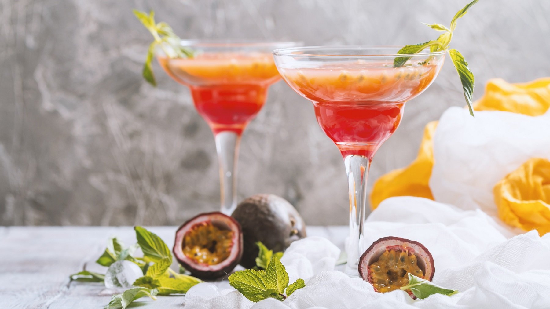Zwei Gläser mit dem Cocktail auf hellgrauem Unter- und dunkelgrauem Hintergrund. Alles dekoriert mit halbierter Passionsfrucht und Minze.