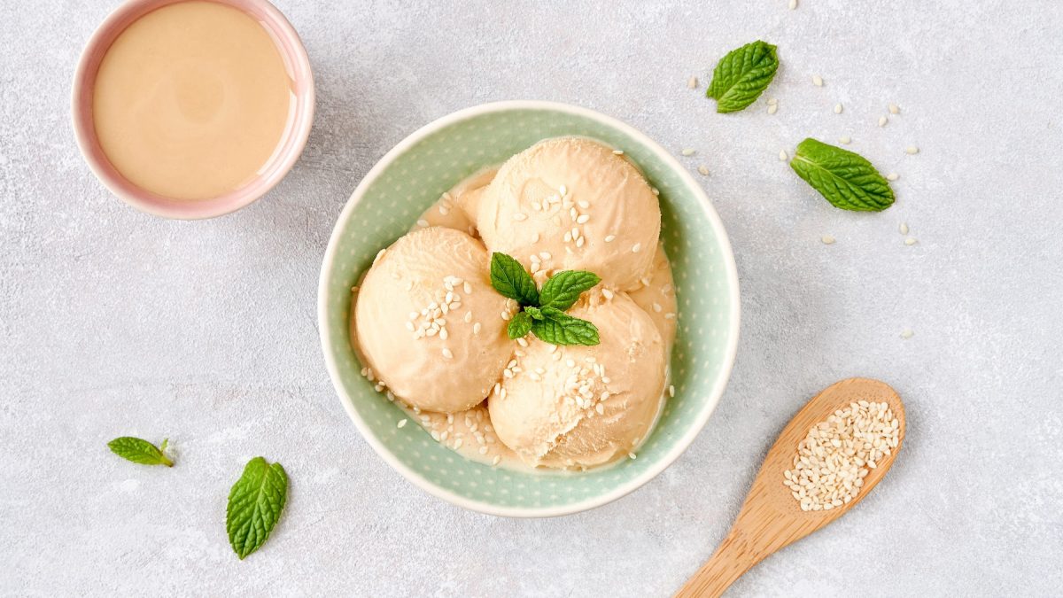 Drei Kugeln Milchmädchen-Eis mit Tahini in einer Keramikschüssel, mit Sesamsamen. Daneben eine Schüssel mit Tahini.