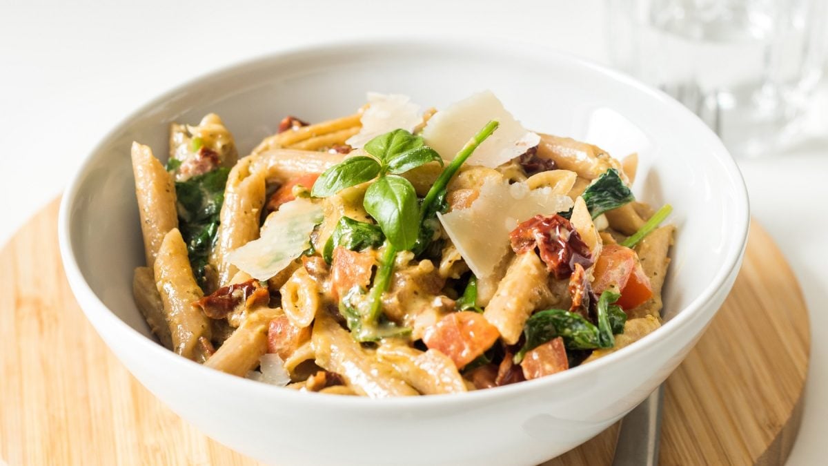 Eine weiße Schale mit dem Nudelsalat auf einem runden Holzbrett und hellem Hintergrund. Alles von leicht schräg oben fotografiert.