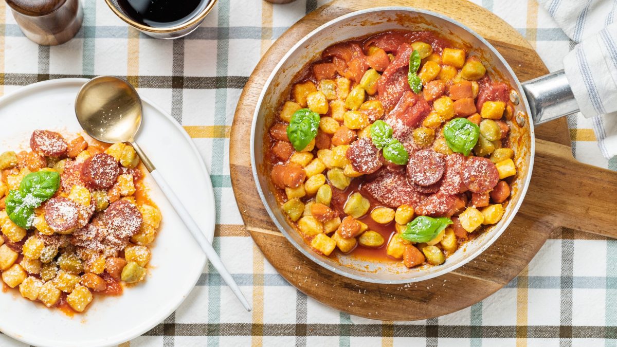 Eine Pfanne und ein weißer Teller mit den Gnocchi auf einem Tisch mit blau-gelb-weiß gestreifter Tischdecke. Danebn noch Salz- und Pfefferstreuer sowie ein Glas mit Rotwein. Alles direkt von oben fotografiert.