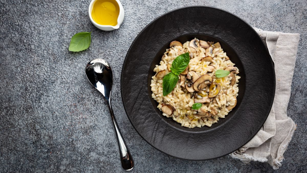 Ein schwarzer Teller mit der One-Pot-Pasta auf grauem Tisch mit Löffel und kleinem weißen Öl-Kännchen. Alles direkt von oben fotografiert.