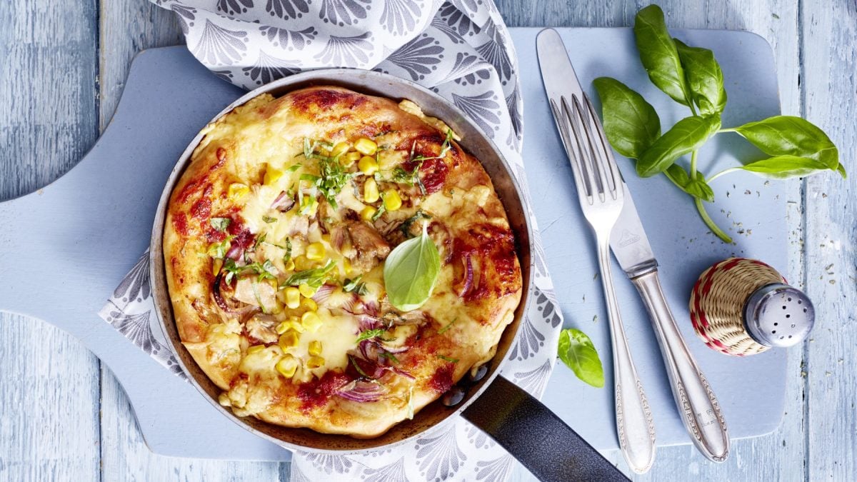 Draufsicht: Pfannen-Pizza mit frischem Basilikum auf in der Pfanne mit hellblauem Untergrund.