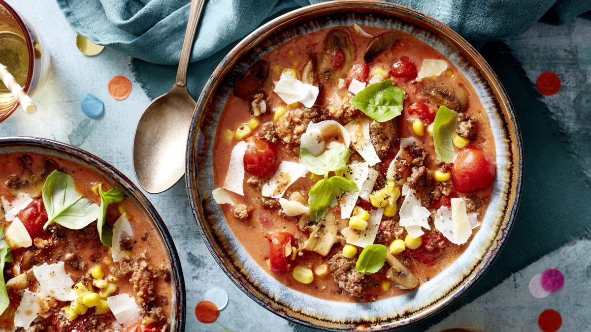 Pizzasuppe mit Hackfleisch, Mais und Gemüse, bestreut mit Käse, auf hellem Steinuntergrund.