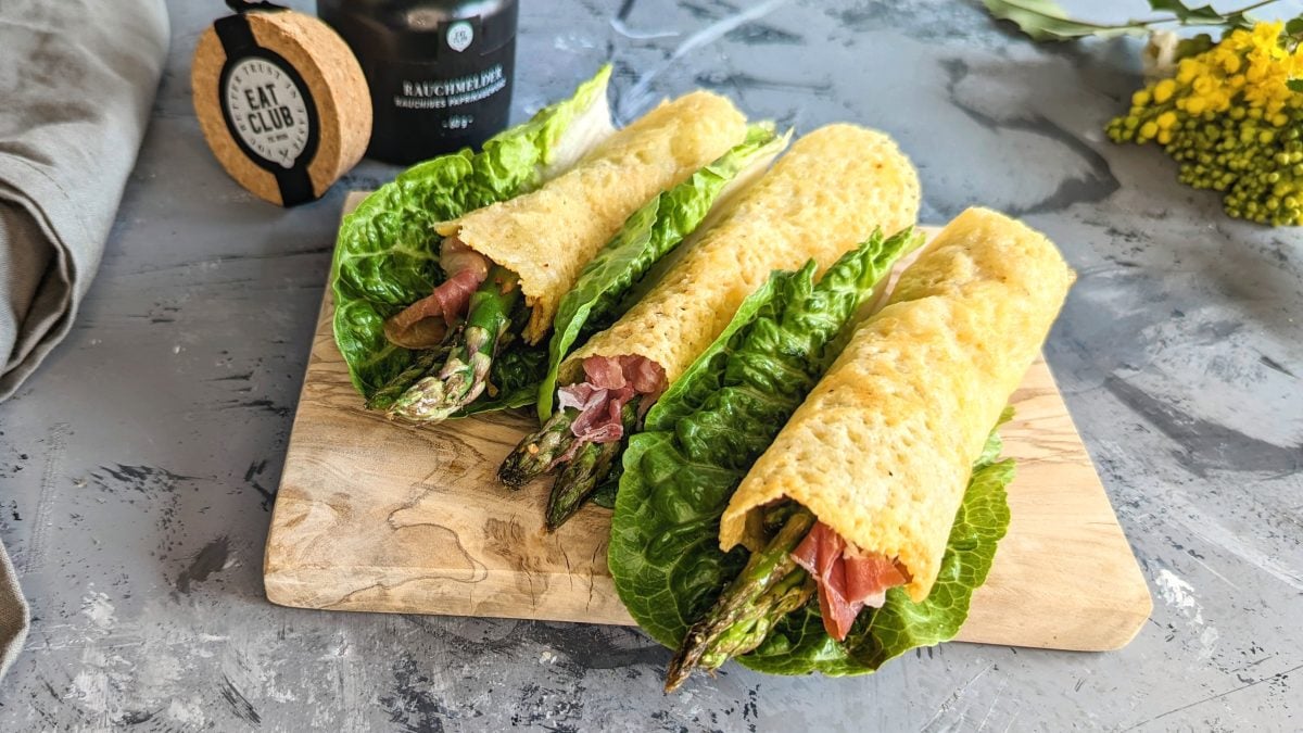 Drei Spargel-Käse-Wraps auf einem kleinen Schneidebrett und grauem Tisch.