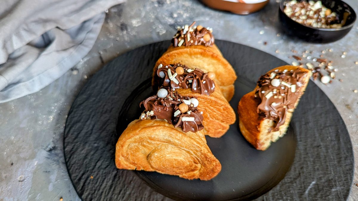 Einige Croissant-Rolls mit Nutella gefüllt auf einem schwarzen Teller und grauem Steinuntergrund. Im Hintergrund eine graue Tischdecke und ein schwarzes Schälchen mit der Schoko-Deko.