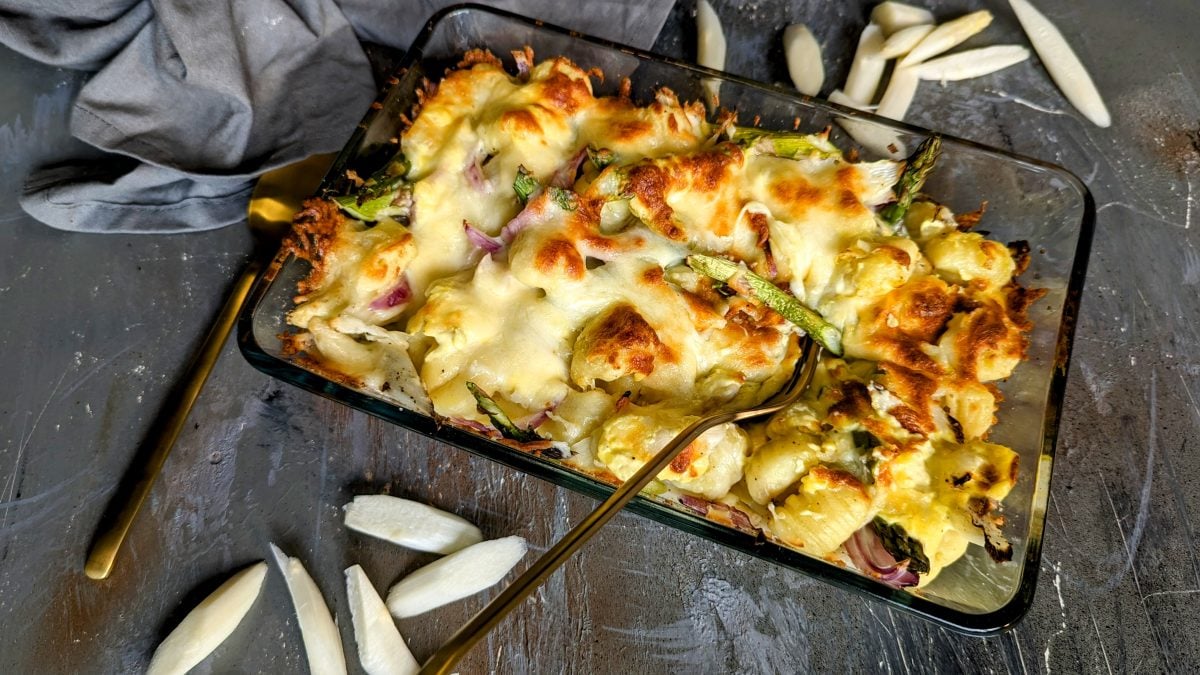 Eine Glasauflaufform mit dem Gnocchi-Gratin mit Spargel und Sauce hollandaise. Alles auf einem grauen Tisch von schräg oben fotografiert.