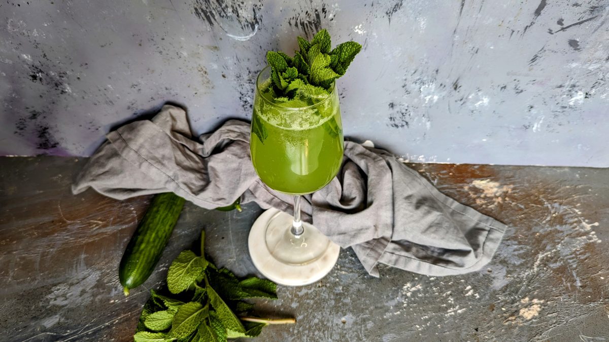 Das Glas mit dem grünen Drink auf grauem Unter- und Hintergrund. Der Tisch ist außerdem mit ein paar Mini-Gurken, Minze und einem grauen Tuch dekoriert.
