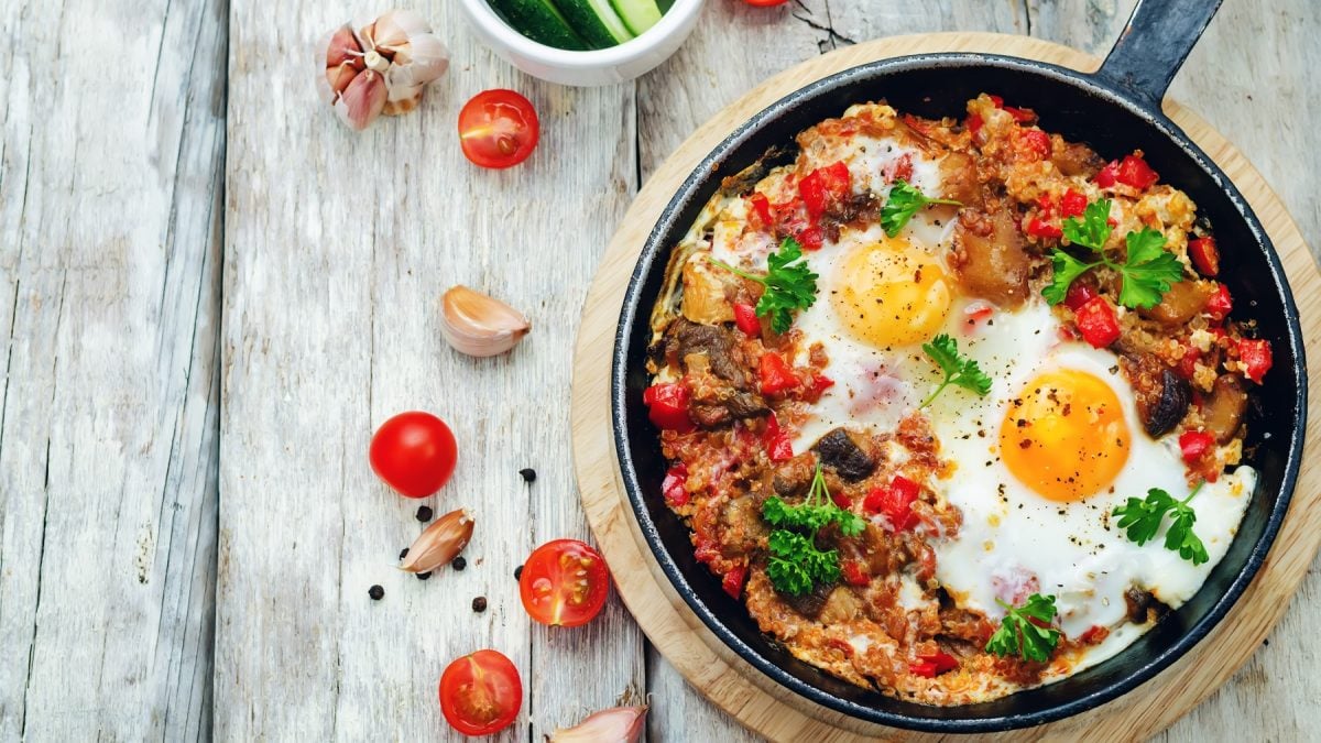 Quinoa-Pfanne mit Gemüse und Eiern in einer Pfanne. Daneben angeschnittene Tomaten und Knoblauchzehen.