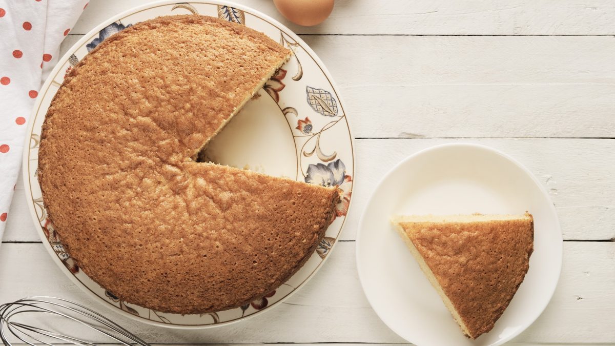 Ein Stück Rührkuchen aus Rührteig auf weißem Teller, neben ganzen Kuchen auf Holztisch.
