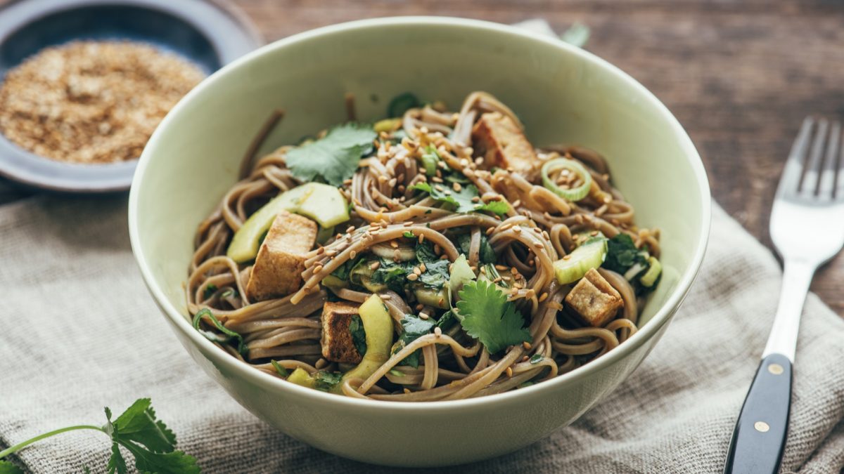 Eine hellgrüne, tiefe Schale mit dem Soba-Salat von schräg oben auf einem Holztisch mit Leinentuch fotografiert.