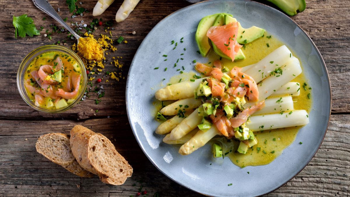 Spargel mit Lachs, Avocado und Zitronensauce auf einem Teller, daneben frisches Brot und eine Schale mit den Hauptkomponenten.