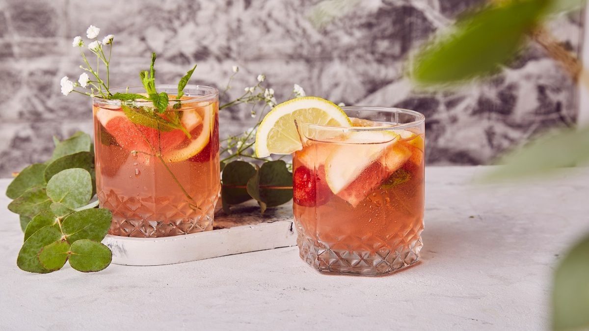 Zwei Gläser mit dem Spring Negroni auf einem hellen Tisch mit grauem Hintergrund. Daneben und dazwischen grüne Pflanzenblätter.