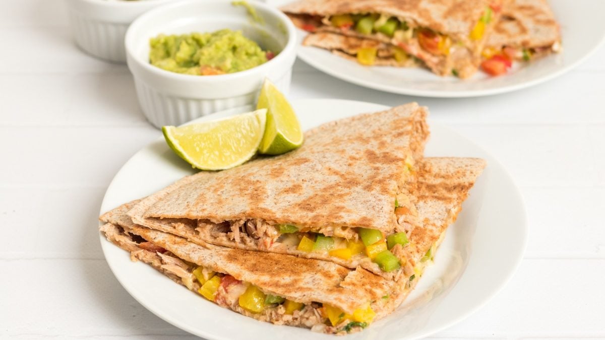 Jeweils 2 Ecken der Quesadilla auf zwei weißen Tellern mit Limettenspalten und einem weißen Schälchen Guacamole. Alles steht auf einem weißen Tisch und ist von der Seite fotografiert.
