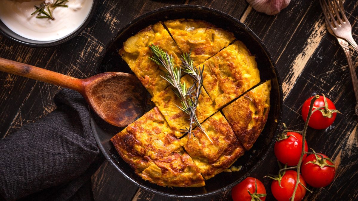 Tortilla de Patatas in einem schwarzen Teller in Stücke geschnitten auf dunklem Hintergrund. Rechts Tomaten und Besteck, links ein Schüsselchen mit einem Dip und ein Löffel.