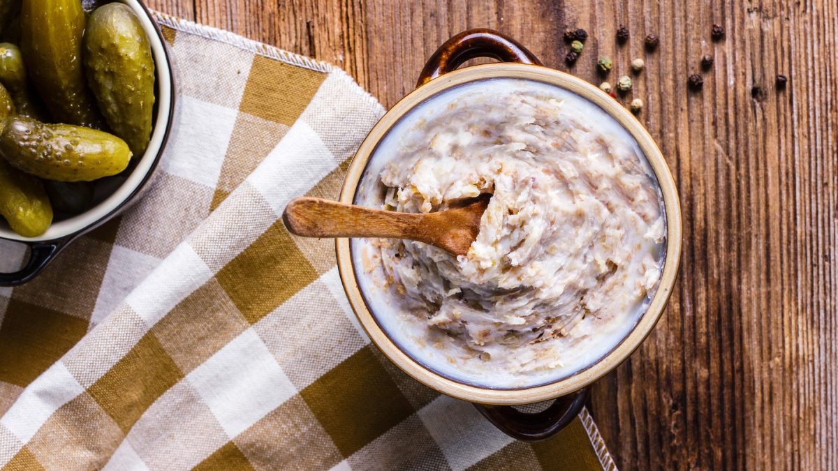 Veganes Zwiebelschmalz in einem Glas mit einem Löffel. Links davon ein Geschirrtuch und ein Glas mit sauren Gurken. Der Hintergrund ist hölzern.