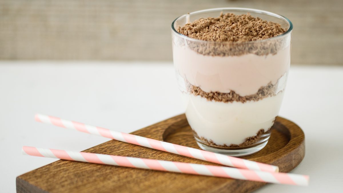 Ein Glas mit dem Dessert und zwei rosa-weißen-Strohhalmen auf einem braunen Untersetzer und hellem Untergrund.